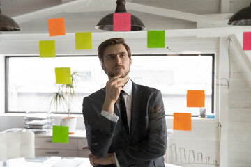 Pensive young Caucasian male employee worker brainstorm develop financial business project on sticky notes in office. Thoughtful businessman plan startup strategy do creative thinking in boardroom.