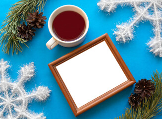 Winter still life. Empty photo frame with space for photo and black tea cup in frame made of pine branches, cones  and decorative snowflakes on blue background. Copy space. Mockup. Flat lay