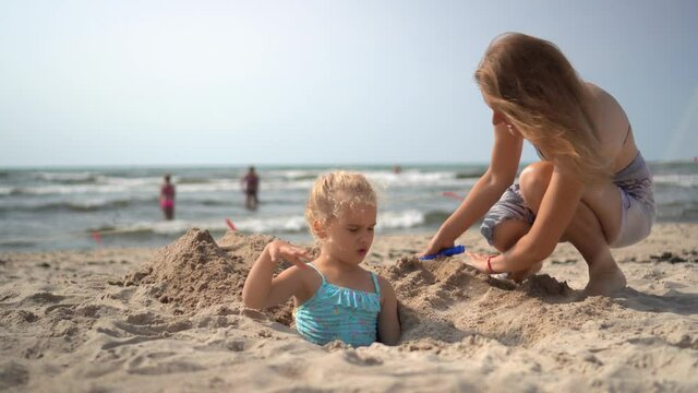 Happy mom bury her little daughter under beach sand. People enjoy summer