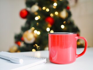 Composition of red mug with coffee, notepad and pen on the white table on the background is a Christmas tree and Christmas lights. Goals 2021. Wish list. Space for text