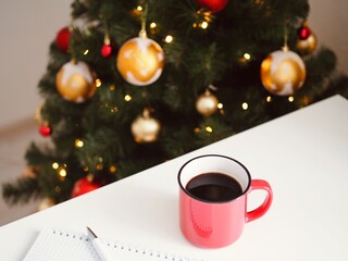 Composition of red mug with coffee, notepad and pen on the white table on the background is a Christmas tree and Christmas lights. Goals 2021. Wish list. Space for text