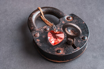 Key to heart concept. Red heart shaped pendant, necklace locked on old rusty lock with key on grey grunge background. Close up. Top view