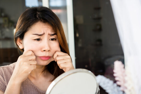 Worried Asian Woman Hand Pulling Her Fat Skin On Cheeks ,checking Her Face On Mirror ,aging And Overweight Concept