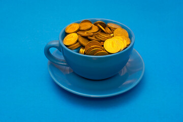 Cheapskate. Blue mug full of shining golden coins on blue saucer against blue background. Love of...
