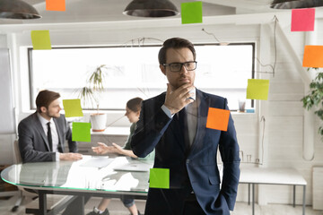 Pensive Caucasian young businessman busy pondering develop business strategy ideas on sticky notes on glass wall. Thoughtful male employee do creative thinking plan or brainstorm in office boardroom.