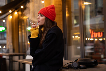 Charming caucasian girl in hat smoking while standing at city street