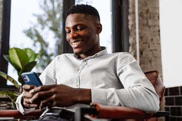 Afro american young man smiling and holding smartphone in armchair