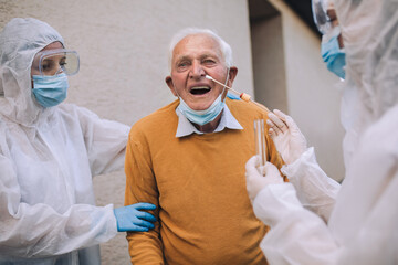 Doctors in a protective suit taking swab from a senior man to test for possible coronavirus infection. Taking corona virus test sample at home concept, senior man in quarantine.