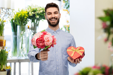 valentine's day, love and people concept - happy smiling man with bunch of peonies and red gift box in shape of heart over flower shop background