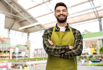 gardening, small business and sale concept - happy smiling male gardener or seller in apron with crossed hands over flower shop background