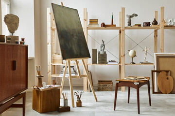 Unique artist workspace interior with stylish teak commode, wooden easel, bookcase, artworks, painting accessories, decoration and elegant personal stuff. Modern work room for artist. Template.