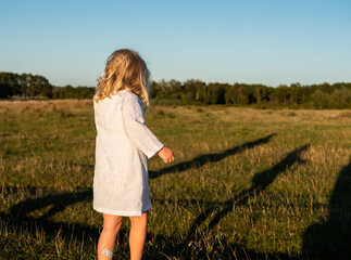 Blond girl 4 years old in white summer dress running and dancing on a field in sunlight and blue sky above