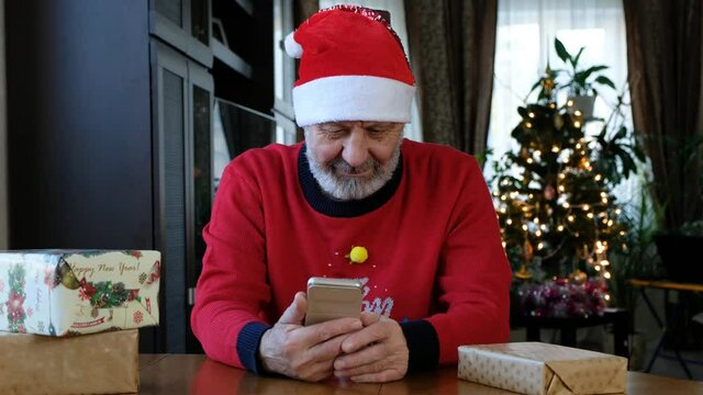 Happy beared senior man in red christmas sweater talking on the smart phone, video call