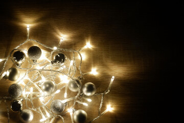 Dark Christmas and New Year composition from garland of yellow led lights and silver shiny, sparkling and matte balls on light oak wood textured surface with copy space