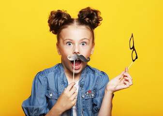 party, childhood and people concept: little girl with a paper accessories over yellow background.
