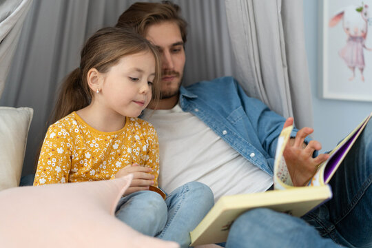 Father reading a book to his daughter while lying on the floor in bedroom.