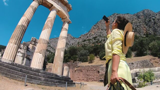 Young woman tourist taking pictures at temple of Athena Pronaia in archaeological site of Delphi, Voiotia, Greece. Fashion elegant yellow dress, large hat, vintage camera.