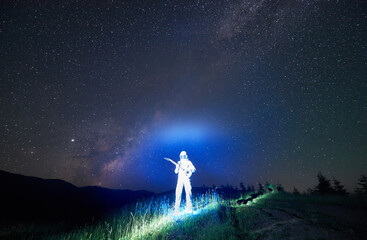 Fototapeta premium Illuminated cosmonaut in white space suit and helmet standing in the middle of mountain meadow holding guitar, incredible night sky full of stars on background. Space travel concept