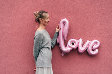 A charming blonde in a gray sweater on a pink photo is holding large inflatable letters LOVE.