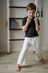 cute caucasian karate boy standing in stance in white kimono pants and black tee shirt at home near wall bars. Individual martial art sport for kids.