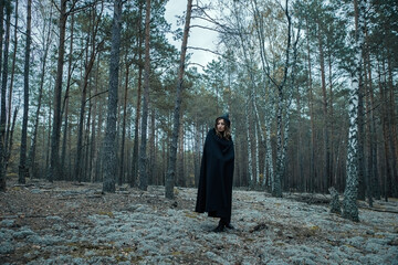 A witch in a black hood is doing magic in the forest.