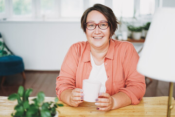 Adult charming brunette woman in glasses plus size body positive with cup of tea at home