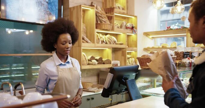 Close up of African American beautiful happy woman seller giving fresh bread baking in bakery shop. Rear of male client paying with credit card on device buying in bakehouse. Food concept