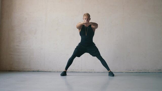 A focused muscular blonde trainer man is doing stretching exercises standing isolated over grey wall