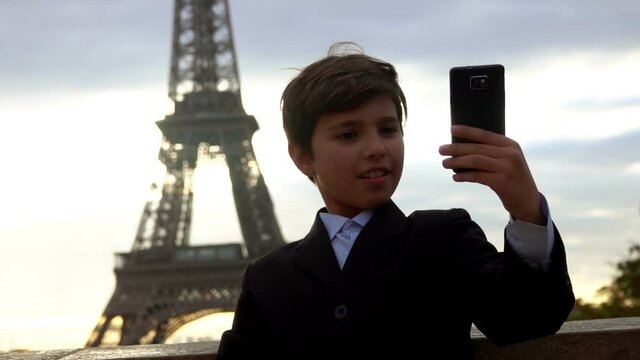 A smiling teenage boy in a black suit is taking a selfie on the phone on the background of the Eiffel tower at the sunrise, Paris, France