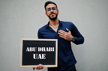 Arab man wear blue shirt and eyeglasses hold board with Abu Dhabi UAE inscription. Largest cities in islamic world concept.