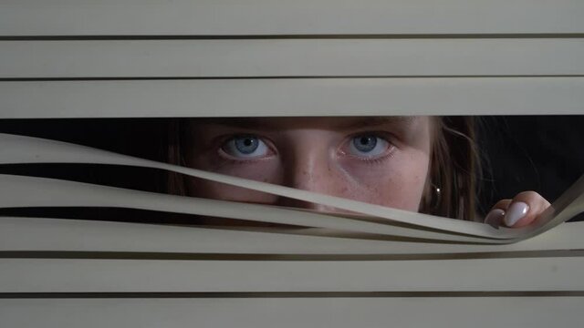 Close up front portrait from indoors of young girl pushing blinds at window with hands. Looking fearfully outside at night