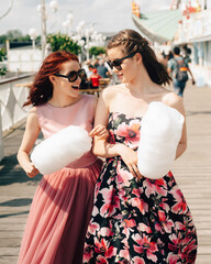 Naklejka premium Close up portrait of smiling excited girls with sunglasses and prom dresses holding cotton candy at amusement park. Teenagers in pink evening dresses on vacation. Birthday party idea.