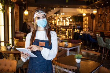 Portrait of female waitress wearing protective face mask while holding touchpad and looking at the camera in a restaurant. Waitress working on touchpad while wearing protective face mask and visor