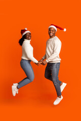 Romantic Black Couple In Santa Hats Holding Hands And Jumping, Orange Background