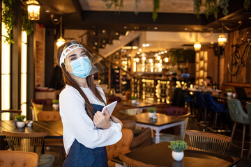 Portrait of a beautiful waitress working at a restaurant wearing a facemask to avoid the spread of...