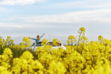 A girl with her hands raised to the sky standing by a car in a yellow flowering field. Freedom and travel concept.