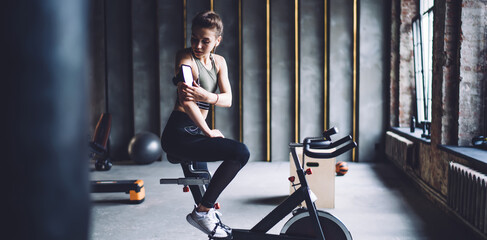 Young sportswoman sitting on cycling machine and fixing smartphone on arm