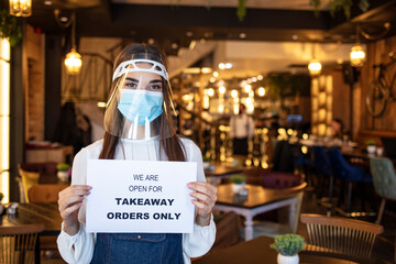 Female waitress hanging open sign while reopening restaurant for delivery and takeaway only during coronavirus epidemic.  Woman holding sign we’re open for takeaway orders only infront of restaurant.