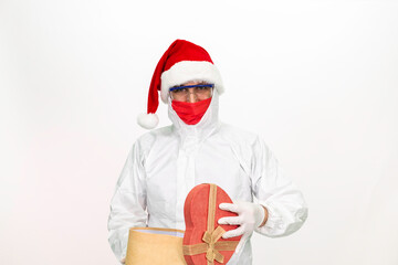 Health worker wearing protective clothing. He wears a santa hat on his head. He has a red corona mask on his face. He is holding a gift box in the shape of a heart.