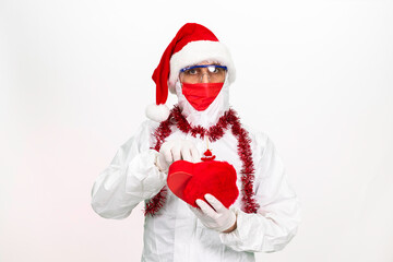 Health worker wearing protective clothing. He wears a santa hat on his head. He has a red corona mask on his face. He is holding a gift box in the shape of a heart.