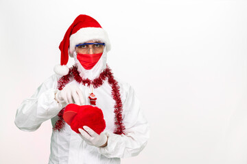 Health worker wearing protective clothing. He wears a santa hat on his head. He has a red corona mask on his face. He is holding a gift box in the shape of a heart.