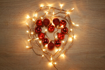 Heart from red shiny, sparkling, matte christmas balls illuminated by a led garland on light oak wood textured surface