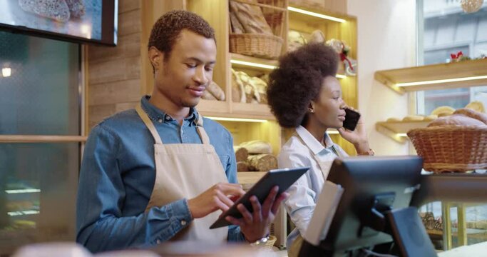 Close up of happy handsome African American man worker in bakehouse typing and tapping on tablet while pretty female coworker speaking on cellphone at work. Entrepreneurs concept