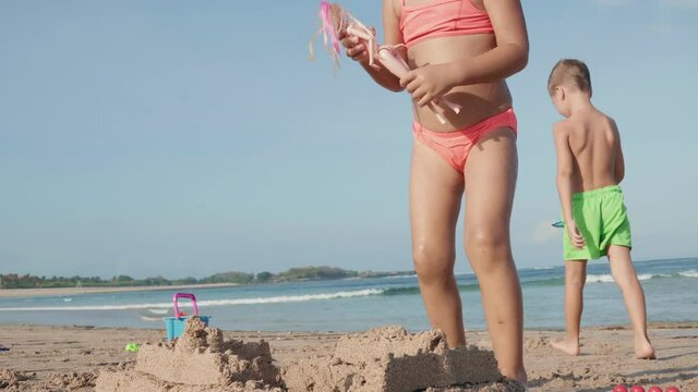 Wide shot of Caucasian boy and his little cute sister wearing swimming suits building sand castle together using toys on the beach