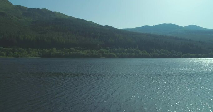 Aerial rising up from grassy banks of Loch Lubnaig in the Trossachs to reveal water and green glen mountains in the background on a sunny day