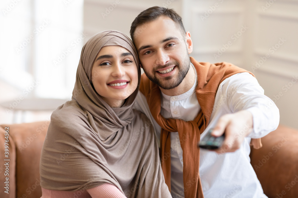 Wall mural young muslim spouses watching television sitting on couch at home