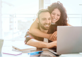 Engaged couple on video call with friends - hugging lovers reading the news in the morning on laptop - warm flare on background.