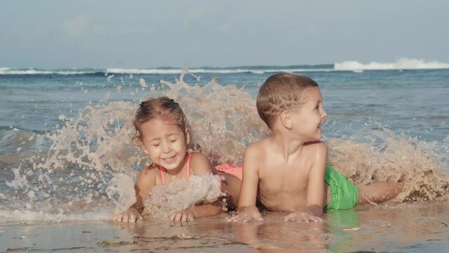 Wide shot of Caucasian boy and his little cute sister wearing swimming suits lying together on sand on seashore and having fun