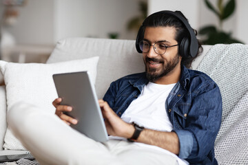 Handsome Arab Guy In Wireless Headphones Relaxing With Digital Tablet At Home
