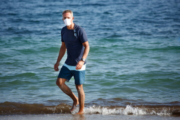 Fototapeta na wymiar Uomo cinquantenne con mascherina protettiva e t-shirt blu sta in spiaggia e passeggia sereno di fronte al mare